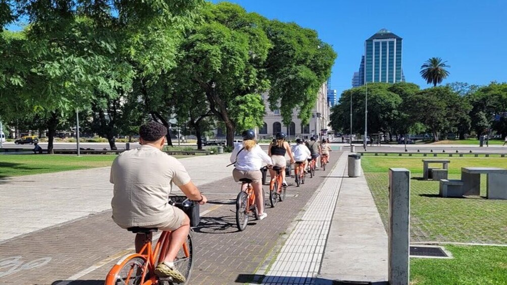 Un grupo de ciclistas en un camino pavimentado en un parque, con árboles y un edificio al fondo. 5