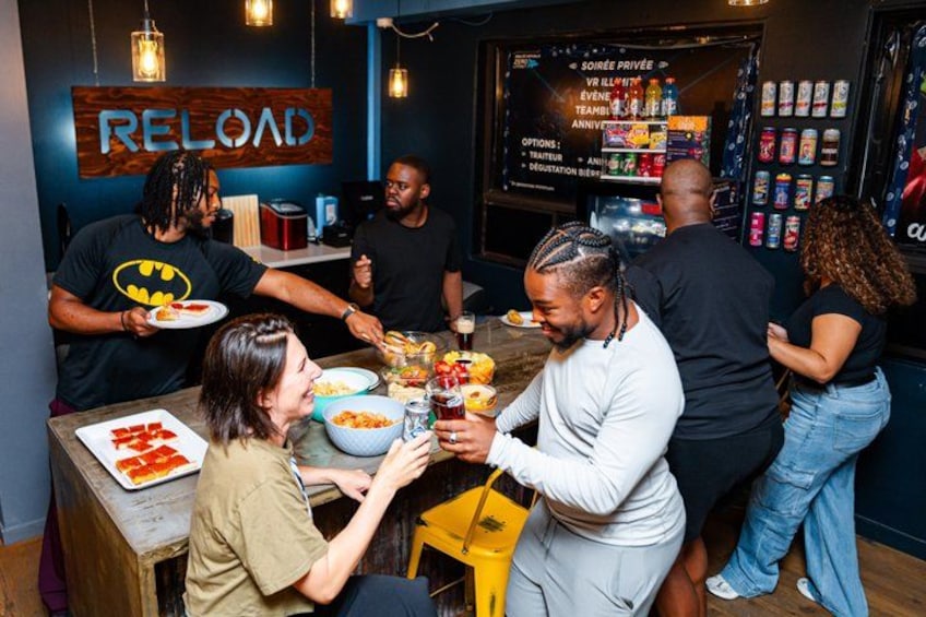 A group of people gathered around a table with food and drinks, in a room with a 'RELOAD' sign on the wall. 4