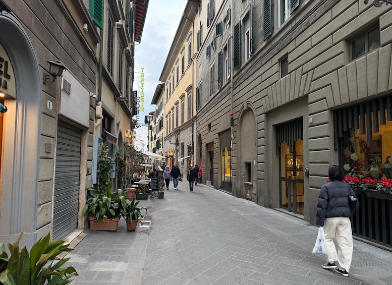 A narrow street lined with buildings, some with shutters down, and a few outdoor seating areas with plants. 4