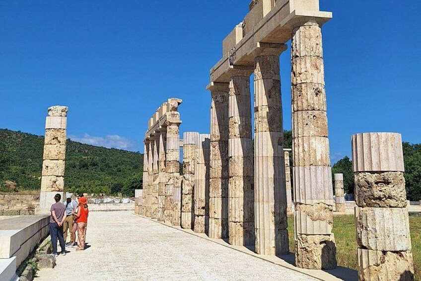 Ancient stone columns, a paved walkway, and a green hillside in the background. 3