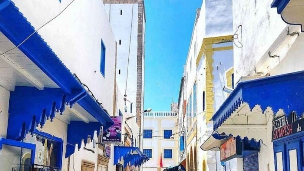 A narrow street with white buildings and blue accents, including awnings and shutters. 3