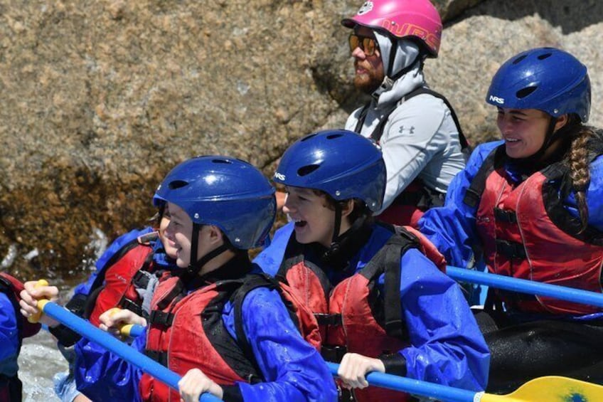 A group of people in a white raft with blue helmets and red life jackets navigating a river. 5