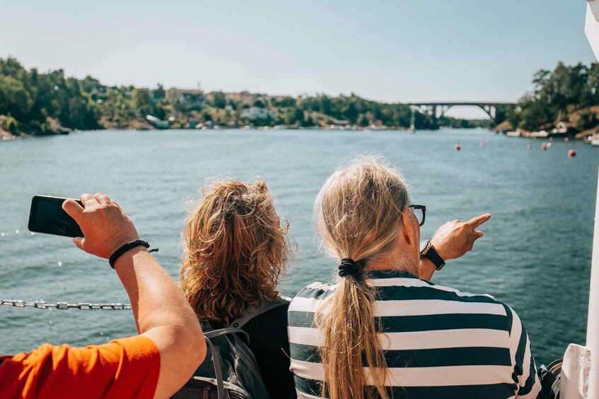 Two people on a boat, one holding a smartphone, with a bridge and trees in the background. 1