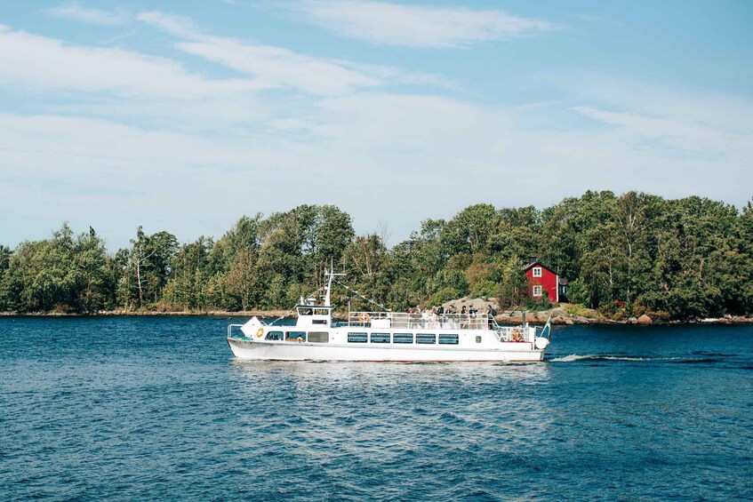 A white boat with passengers on a calm body of water, surrounded by a forested shoreline with a red building. 4