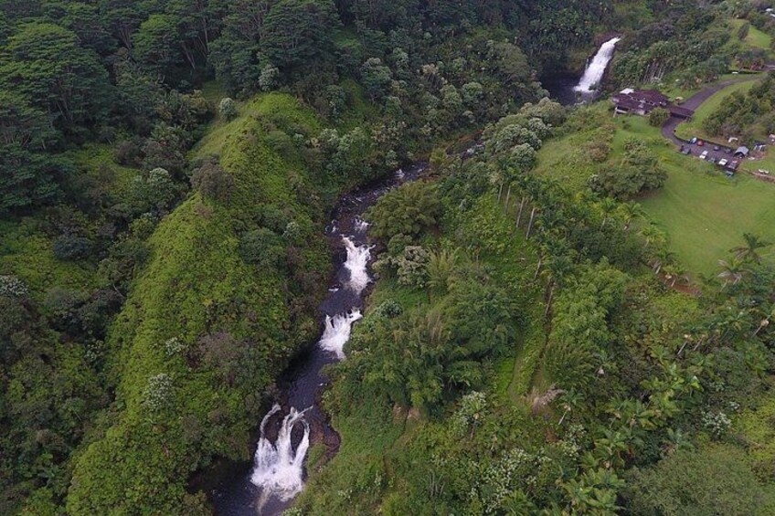 Ein Wasserfall stürzt durch ein üppiges, grünes Tal, in dessen Nähe sich ein kleines Gebäude und ein Parkplatz befinden. 2