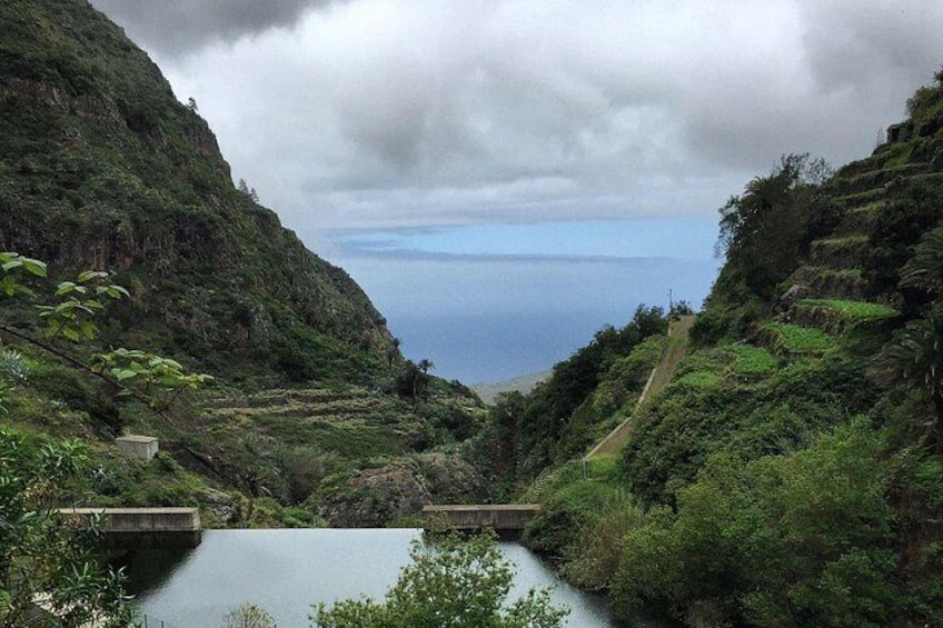A mountainous landscape with a river, a bridge, and a view of the ocean under cloudy skies. 3