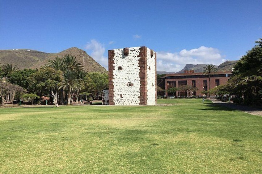 A tower with a brick pattern, a grassy field, and a building with a red roof in the background. 4