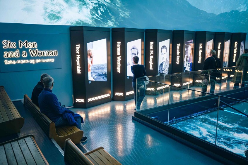 A museum exhibit with a large wall displaying portraits and names of individuals, benches, and a glass railing overlooking a water feature. 5