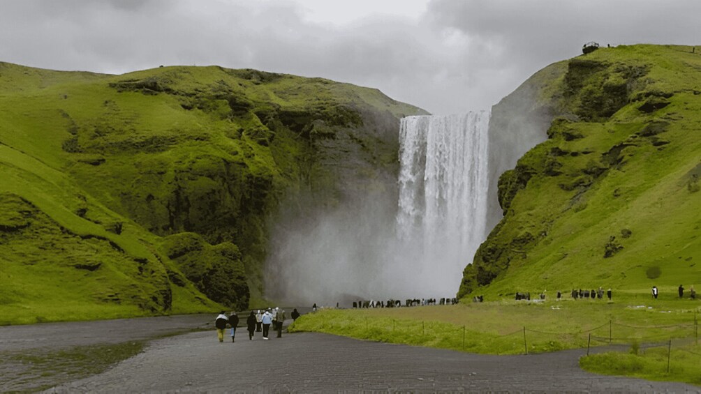 A waterfall surrounded by green hills with people walking in the foreground. 2