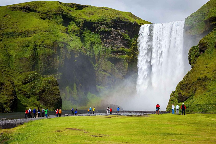 A waterfall surrounded by lush green hills with people standing on a path in front. 1
