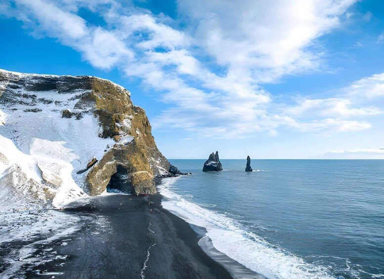 A snowy beach with a large rock formation and two smaller rocks in the water. 5