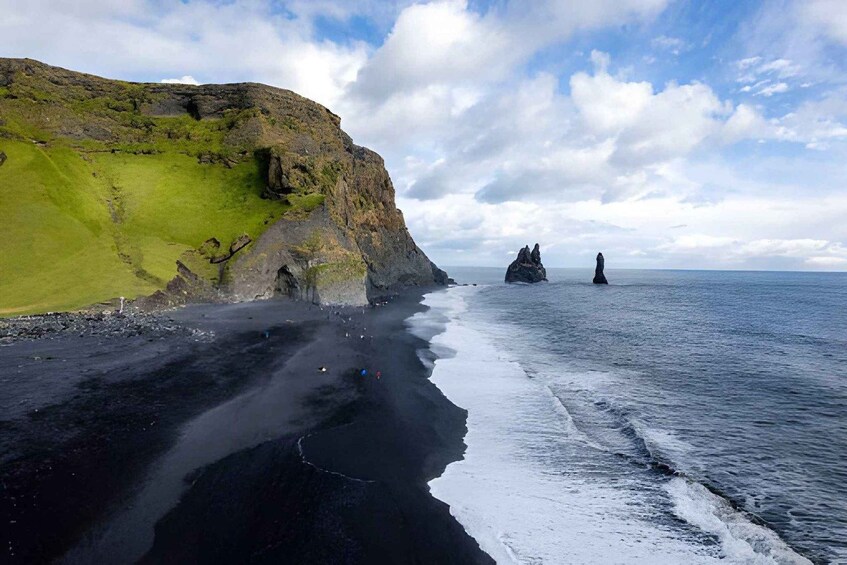A coastal landscape with a black sand beach, green grassy cliffs, and two prominent rock formations in the sea. 4