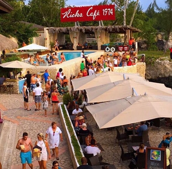 A crowded outdoor café with a pool and umbrellas. 4