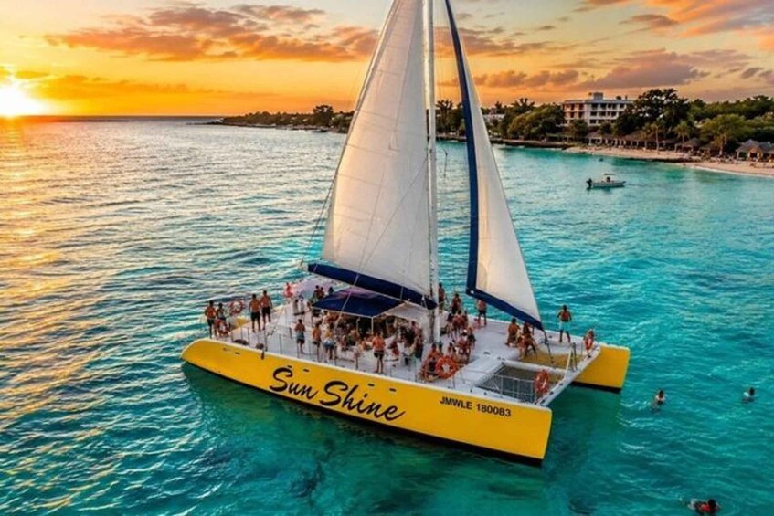 Eine Segelyacht namens 'Sun Shine' mit Personen an Bord, segelt bei Sonnenuntergang über türkisfarbenes Wasser. 1