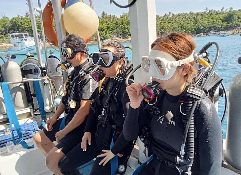 Three people in scuba diving gear on a boat, with a tropical beach and water in the background. 4