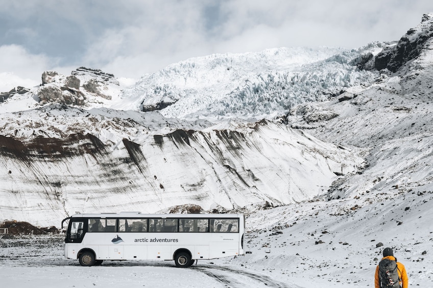 A white bus with 'arctic adventures' written on it, parked on a snow-covered road with a mountainous landscape in the background. 3