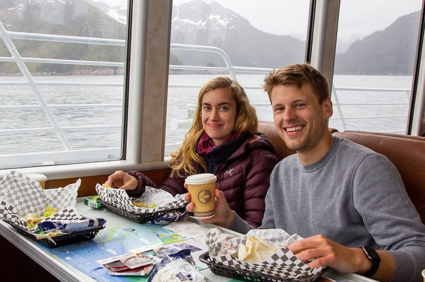 To personer sidder ved et bord med mad og drikke på en båd. 4