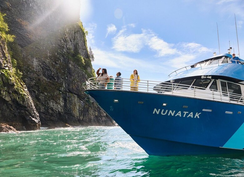 A blue boat named NUNATAK with passengers on deck, surrounded by clear turquoise water and rocky cliffs. 1