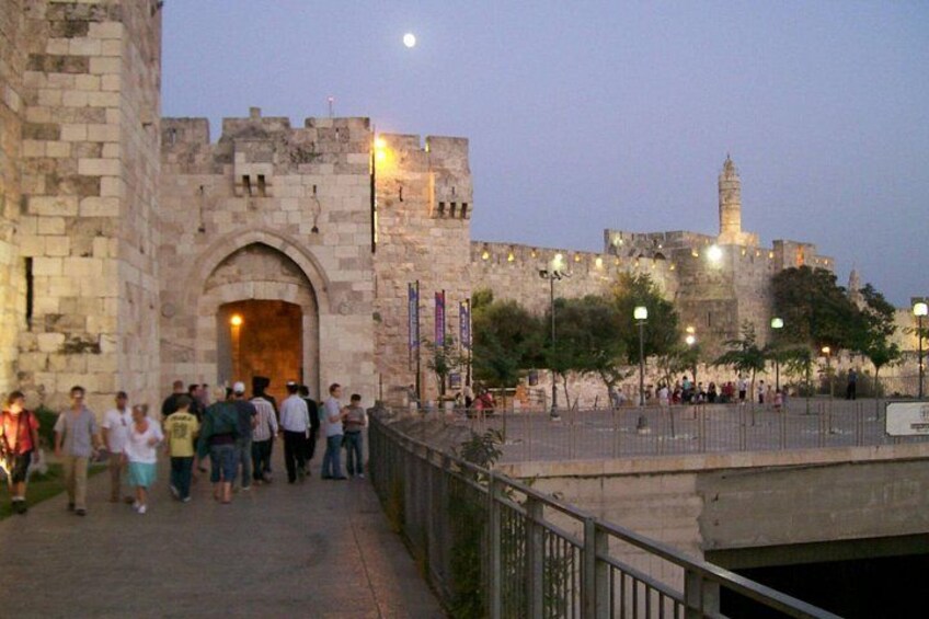 A historic stone gate with people walking in front, a large building with a tower in the background, and street lamps along the pathway. 4