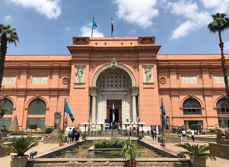 A large, pink building with a central archway and statues on either side. There are flags on top and people walking in front. 4