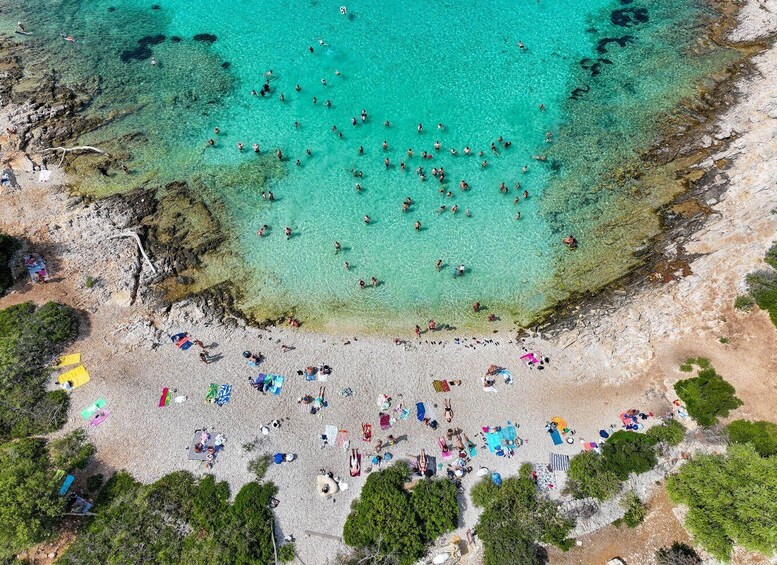 An aerial view of a beach with clear turquoise water, people swimming and sunbathing, and scattered umbrellas. 1