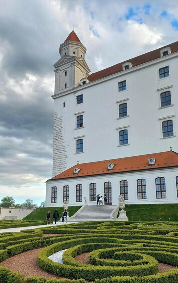 Un grand bâtiment blanc avec un toit rouge et une tour. On peut y voir des statues devant le bâtiment, ainsi qu’un jardin bien entretenu avec des haies. 5