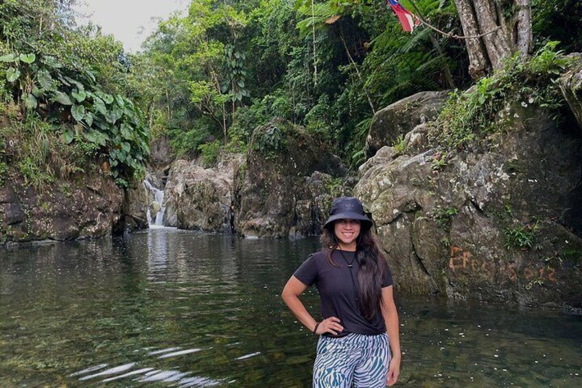 Una persona de pie junto a un arroyo cristalino con una cascada al fondo, rodeada de abundante vegetación. 2