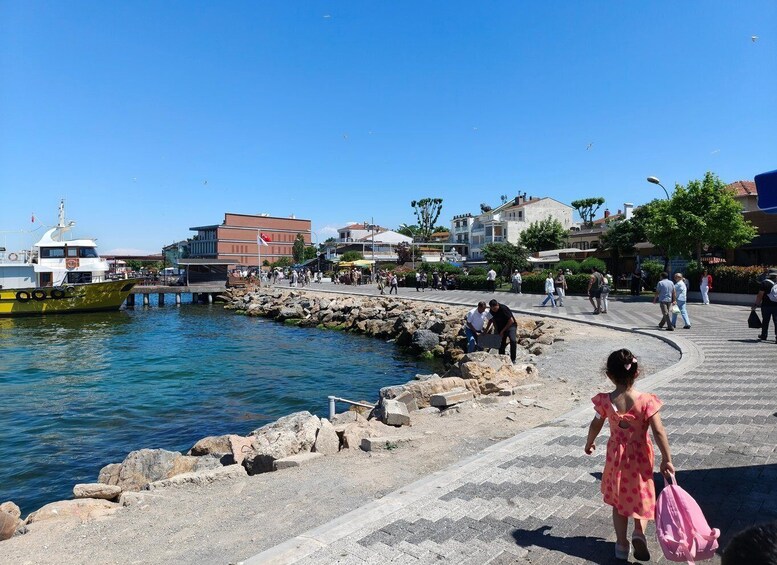 A coastal promenade with people walking, a yellow boat docked, and buildings in the background. 3