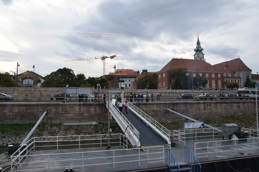 A bridge over a river with a construction crane in the background, a red building with a clock tower, and several parked cars. 2
