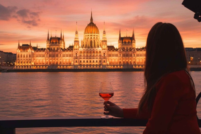 A person holding a glass of red wine, overlooking a grand building with a dome and spires, by a body of water during sunset. 1
