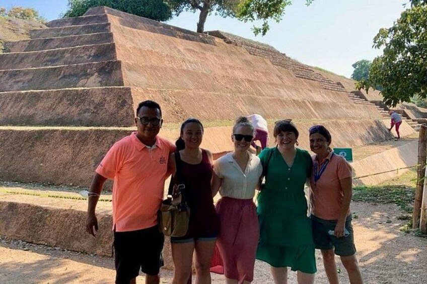 A group of people standing in front of a large, stepped structure with a tree in the background. 4