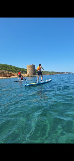 Two people paddleboarding on a clear blue sea with a historic tower in the background. 3