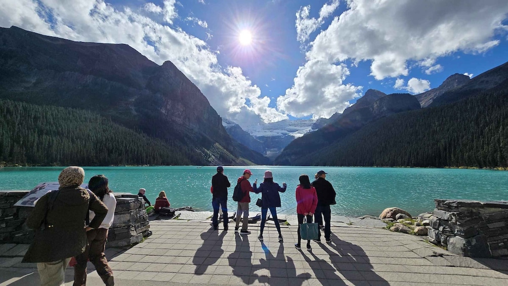 A group of people standing on a dock overlooking a turquoise lake with mountains in the background. 2