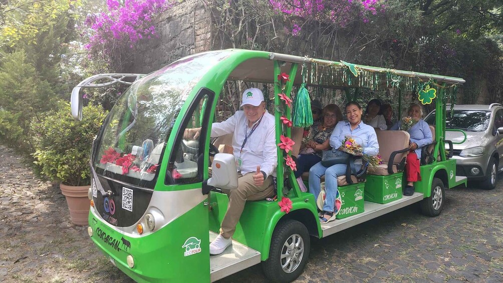 A green tour bus with passengers, parked on a cobblestone path surrounded by greenery and flowers. 5