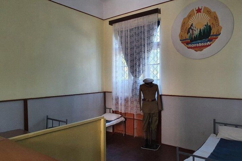 Una habitación con cama, una ventana con cortinas, un maniquí y una pared con un escudo de armas. 2