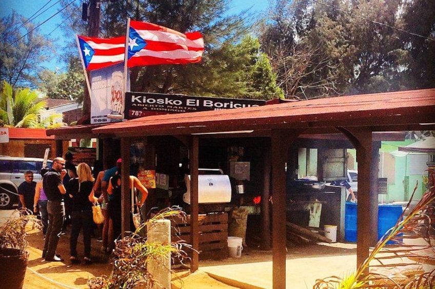 A small outdoor café named Kiosk El Boricua with a Puerto Rican flag, a signboard, and a few people standing outside. 1