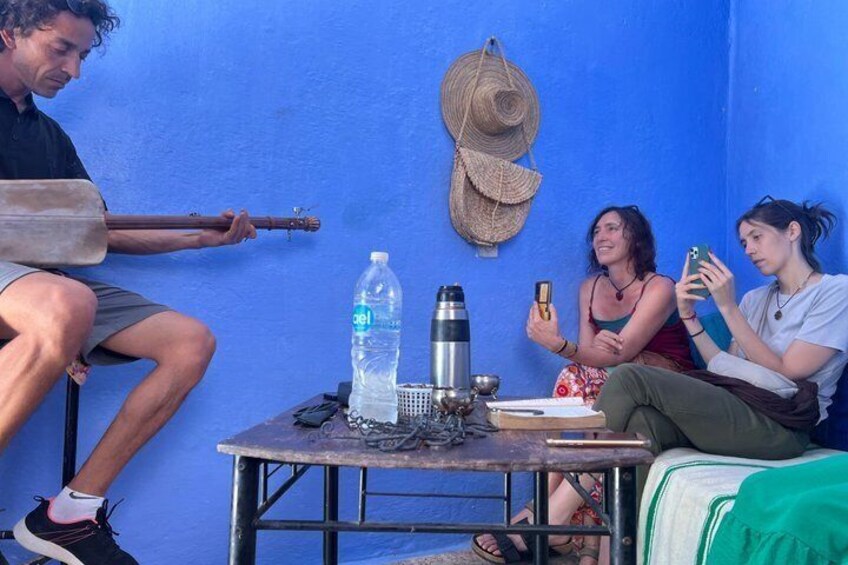 Un groupe de personnes est assis autour d’une table. On aperçoit une guitare, une bouteille d’eau et un chapeau accroché au mur. 2