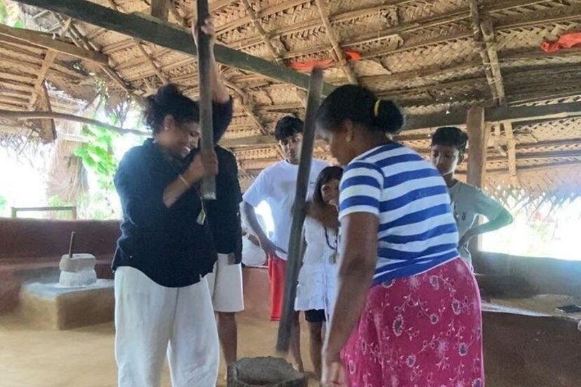 A group of people under a thatched roof structure, engaged in an activity involving a wooden tool. 4