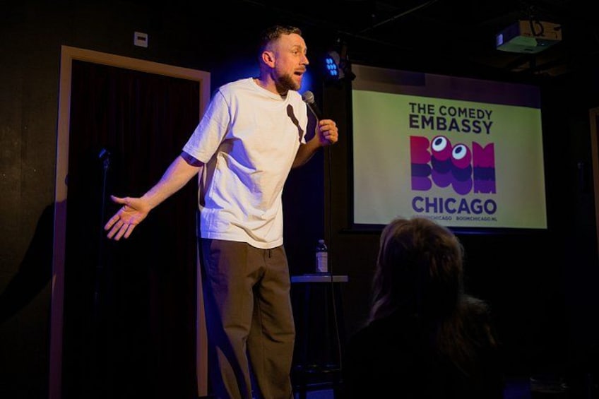 Una persona de pie en un escenario, un micrófono, una pantalla mostrando el texto 'The Comedy Embassy Boom Chicago' y una puerta al fondo. 1