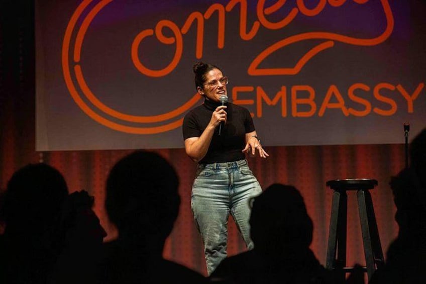 Una persona hablando en un escenario con un micrófono, un taburete y un fondo con el texto 'Embassy'. 5