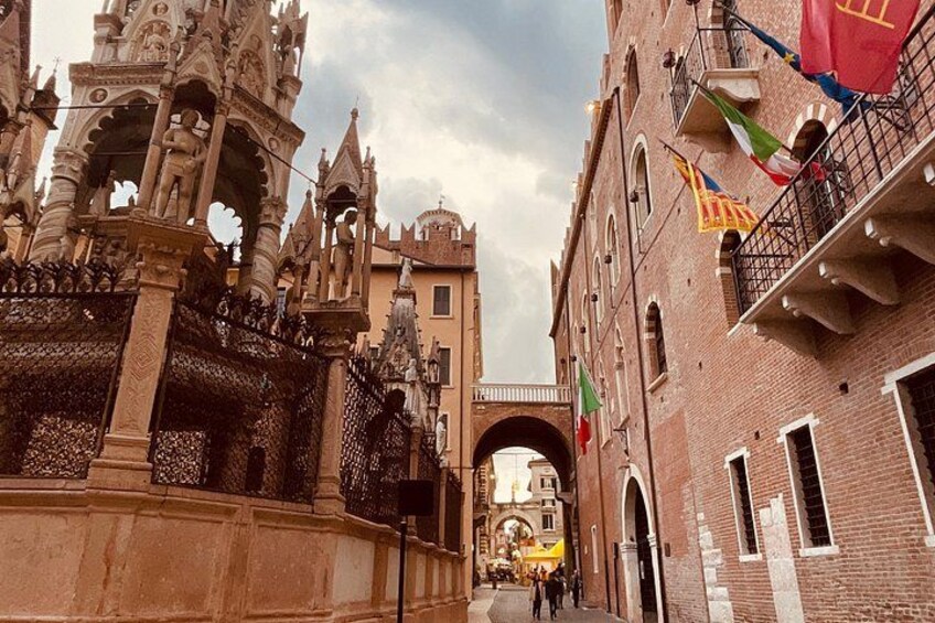A historic street with ornate architecture, including a Gothic-style gate and a brick building with flags. 3
