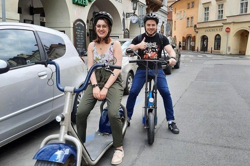 Two people on electric scooters, a car, and a street with buildings in the background. 5