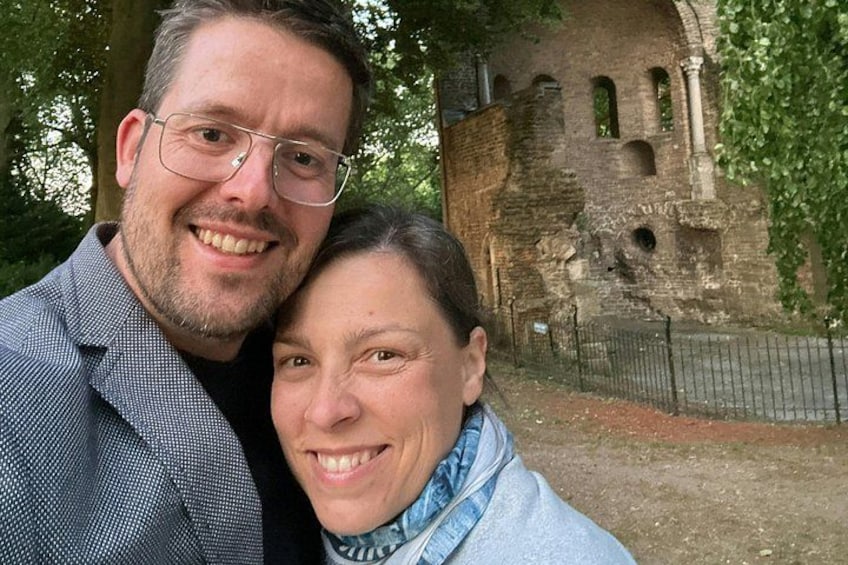 Et par står foran en historisk stengbygning med buer og et hegn. 2