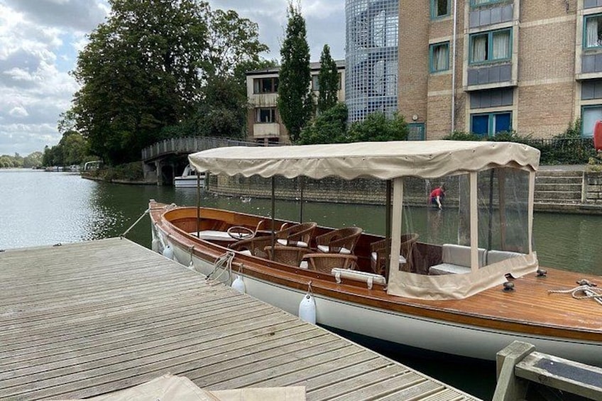 Un bote de madera amarrado en un muelle con una zona de estar techada, un puente al fondo y edificios a lo largo de la orilla del río. 4