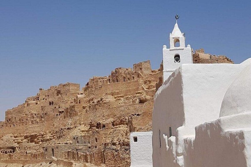 A white building with a bell tower, a rocky hill with ruins, and a clear blue sky. 5