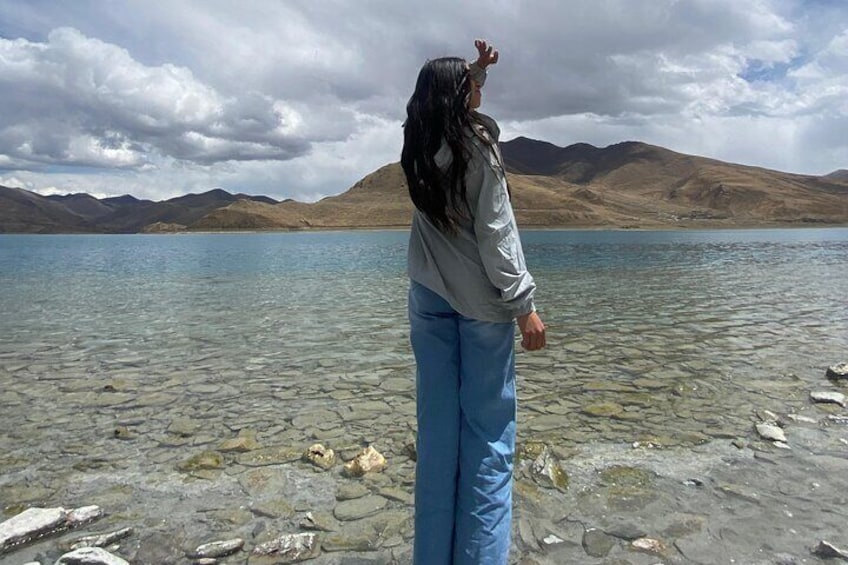 A person standing by a clear lake with rocky shore, mountains in the background, and a cloudy sky. 3