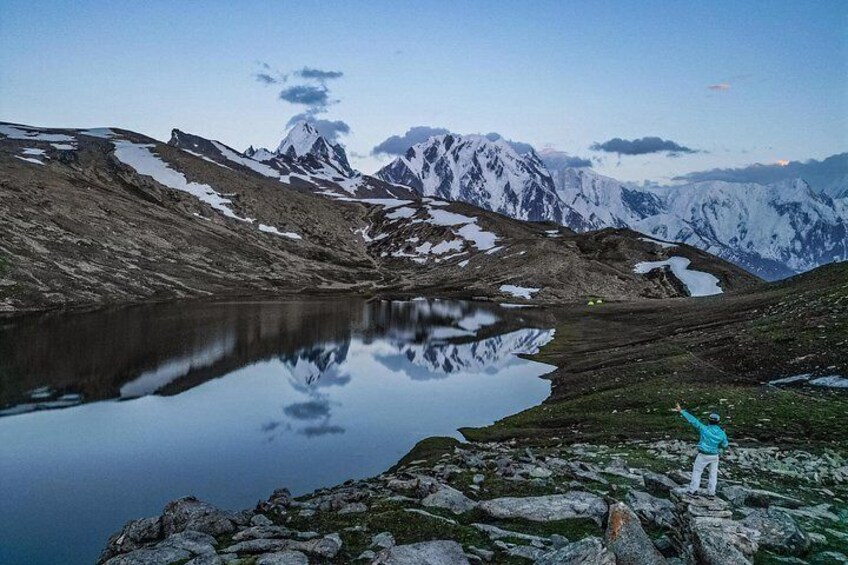 Un paisaje montañoso con un lago, una persona de pie en la orilla y picos nevados al fondo. 2
