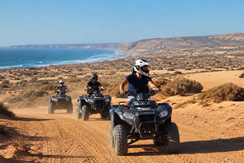 Three people riding ATVs on a sandy path with a desert landscape and ocean in the background. 1