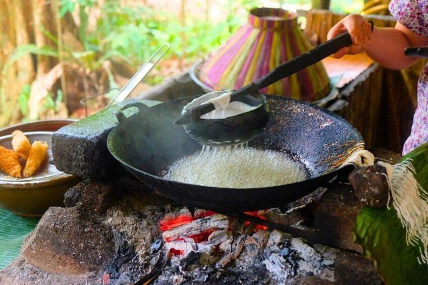En matlagningsaktivitet utomhus med en stekpanna på ett spris, en person som rör om i innehållet och en traditionell kastrull i bakgrunden. 3