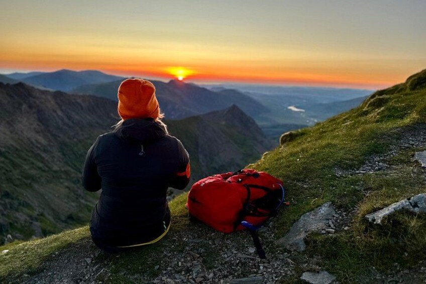 A person sitting on a mountain path with a red backpack, overlooking a valley during sunset. 1